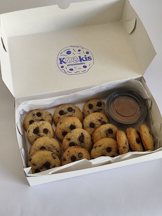 Caja blanca con el sello de Kookis abierta, llena de galletas con chips y un pote de dulce de leche para mojar.